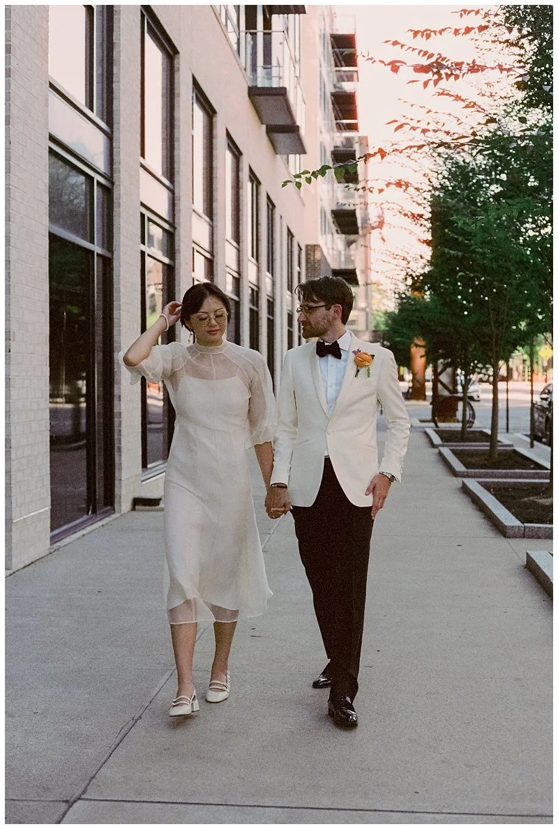 A couple dressed in wedding attire walking hand in hand on a sidewalk in an urban setting during sunset.