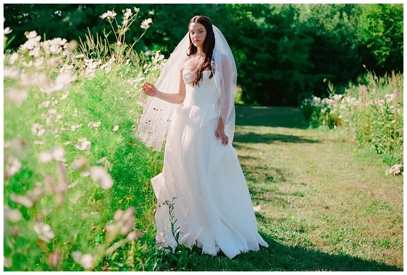A bride in a white wedding dress and veil standing in a lush green garden with pink flowers and trees in the background.