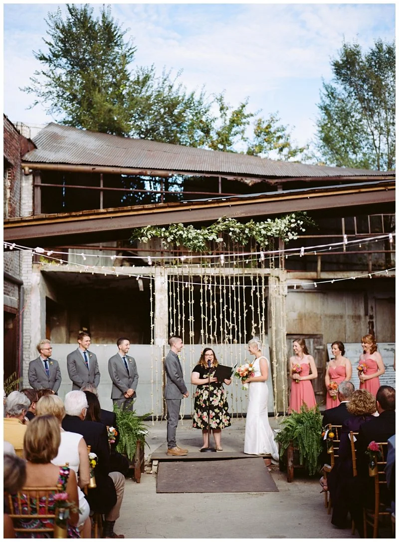 Couple exchanging vows at an outdoor wedding ceremony with guests seated around, bridesmaids in pink dresses, groomsmen in suits, and floral decorations in an industrial setting.