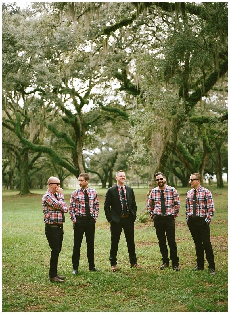 Five men standing on grass in a wooded area, dressed in matching plaid shirts, with one man in a suit, smiling and talking. Large trees with hanging moss surround them.