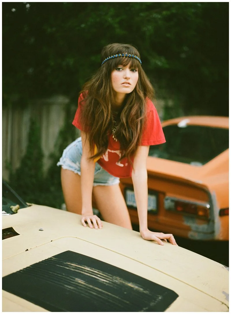 A young woman with long wavy hair wearing a red t-shirt, denim shorts, and a blue headband, leaning forward on a yellow car with a black stripe on the hood. There is an orange car and greenery in the background.