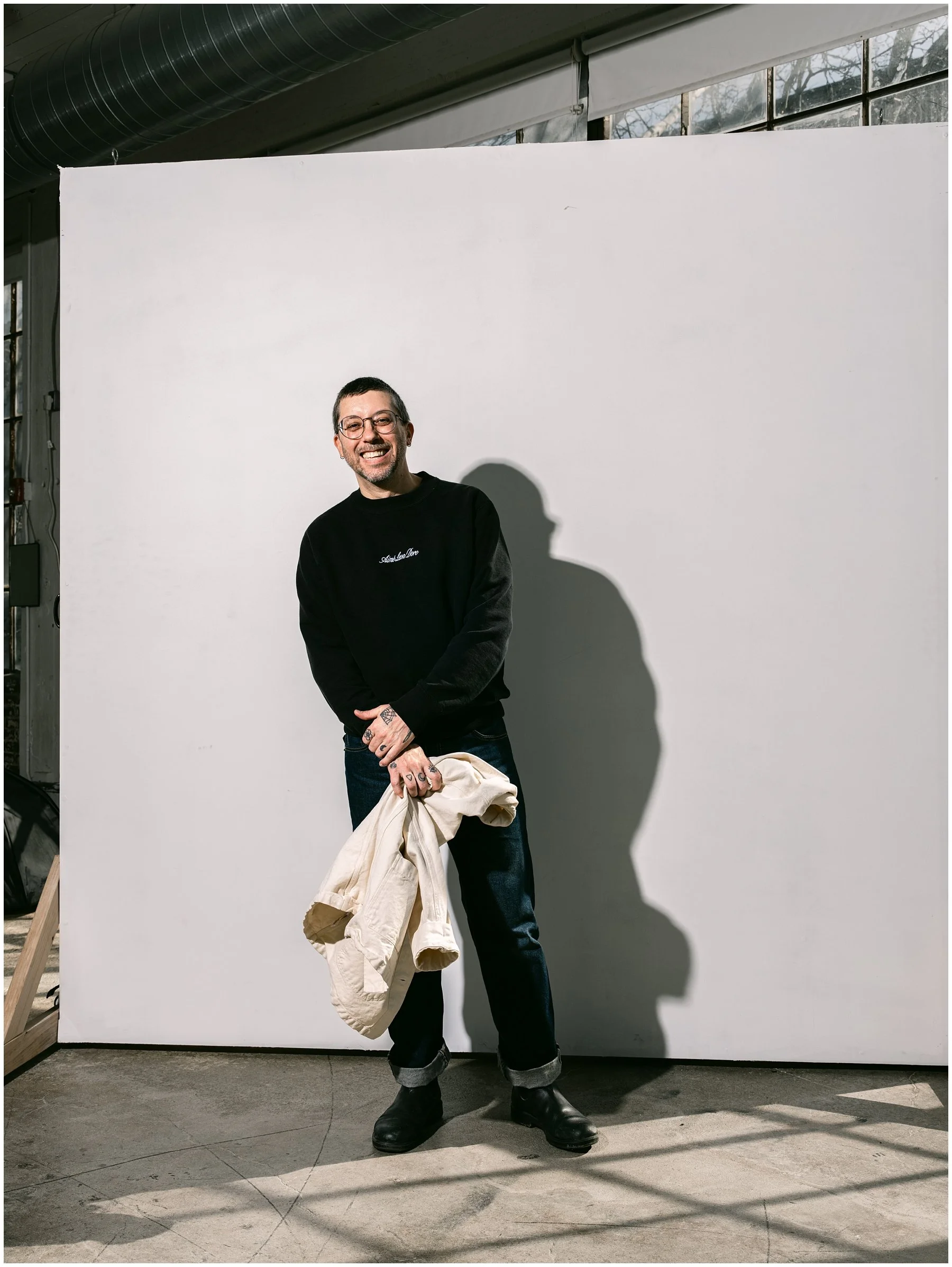 A man with glasses, dark hair, and a beard, smiling stands in front of a plain white background, holding a light-colored jacket, dressed in a black sweater, dark jeans, and black boots. Adam Lowe in his studio
