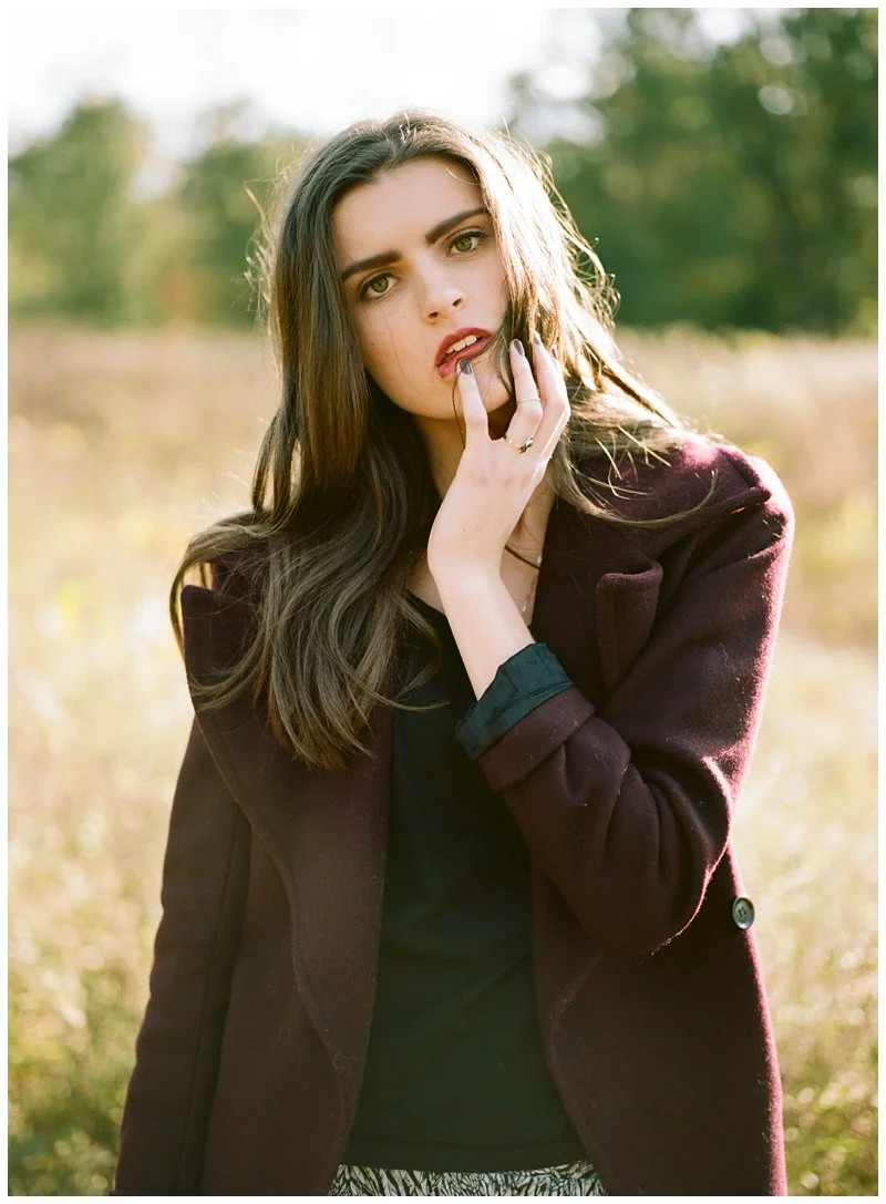 A young woman with long dark hair and fair skin outdoors, wearing a maroon coat over a black top, with a hand touching her face, in a natural setting with trees and sunlight. Shot on film