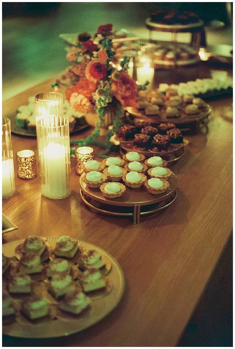 A table decorated with lit candles and a floral centerpiece, set with various plates of desserts including cookies, cupcakes, and other treats.
