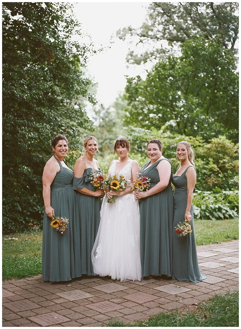 A bride and four bridesmaids standing outdoors on a brick pathway surrounded by greenery, holding bouquets of flowers, smiling for a group photo.