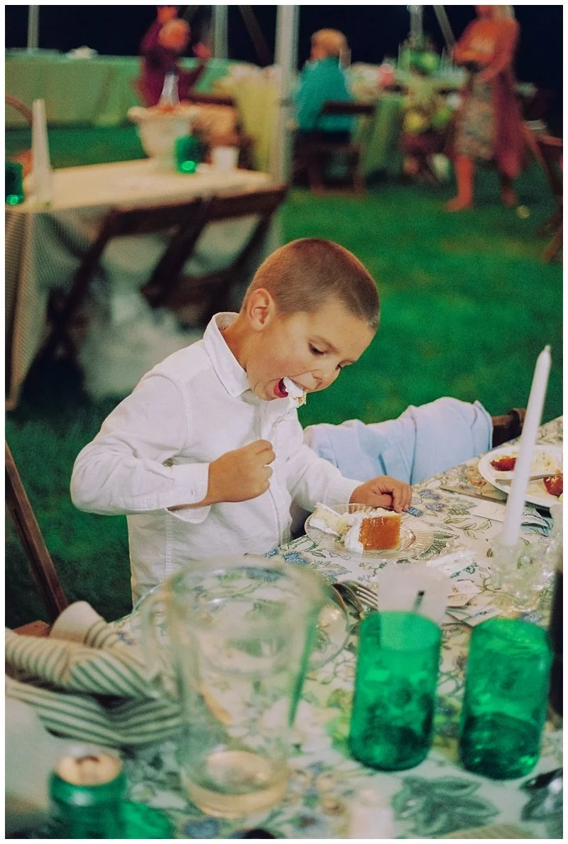 A young boy in a white shirt is sitting at a table, eating cake at an outdoor gathering or party. The table has a floral tablecloth, green glasses, and various dishes. In the background, there are other people and party decorations.