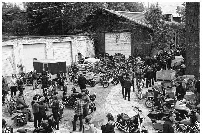 Crowd of people gathered in a lot filled with motorcycles and bikes, with old buildings and trees in the background.