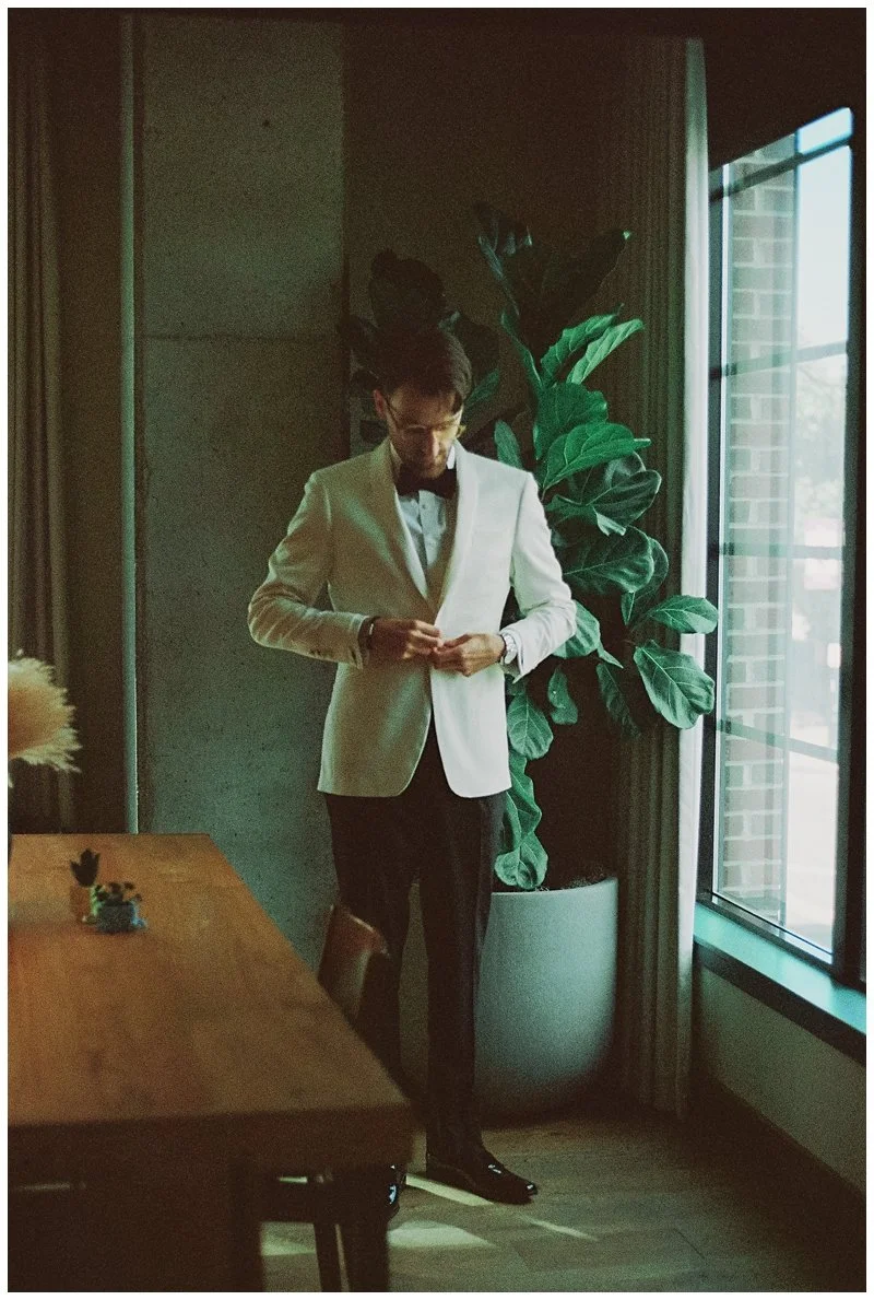 A man dressed in a white tuxedo jacket, black pants, and a black bow tie standing near a large plant in a room with a window.