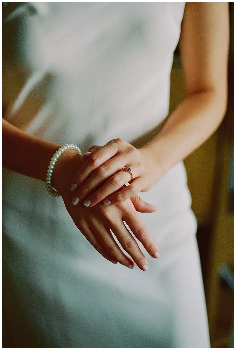 Close-up of a woman wearing a white dress, with a pearl bracelet and a wedding ring on her left hand, kissing her right hand.