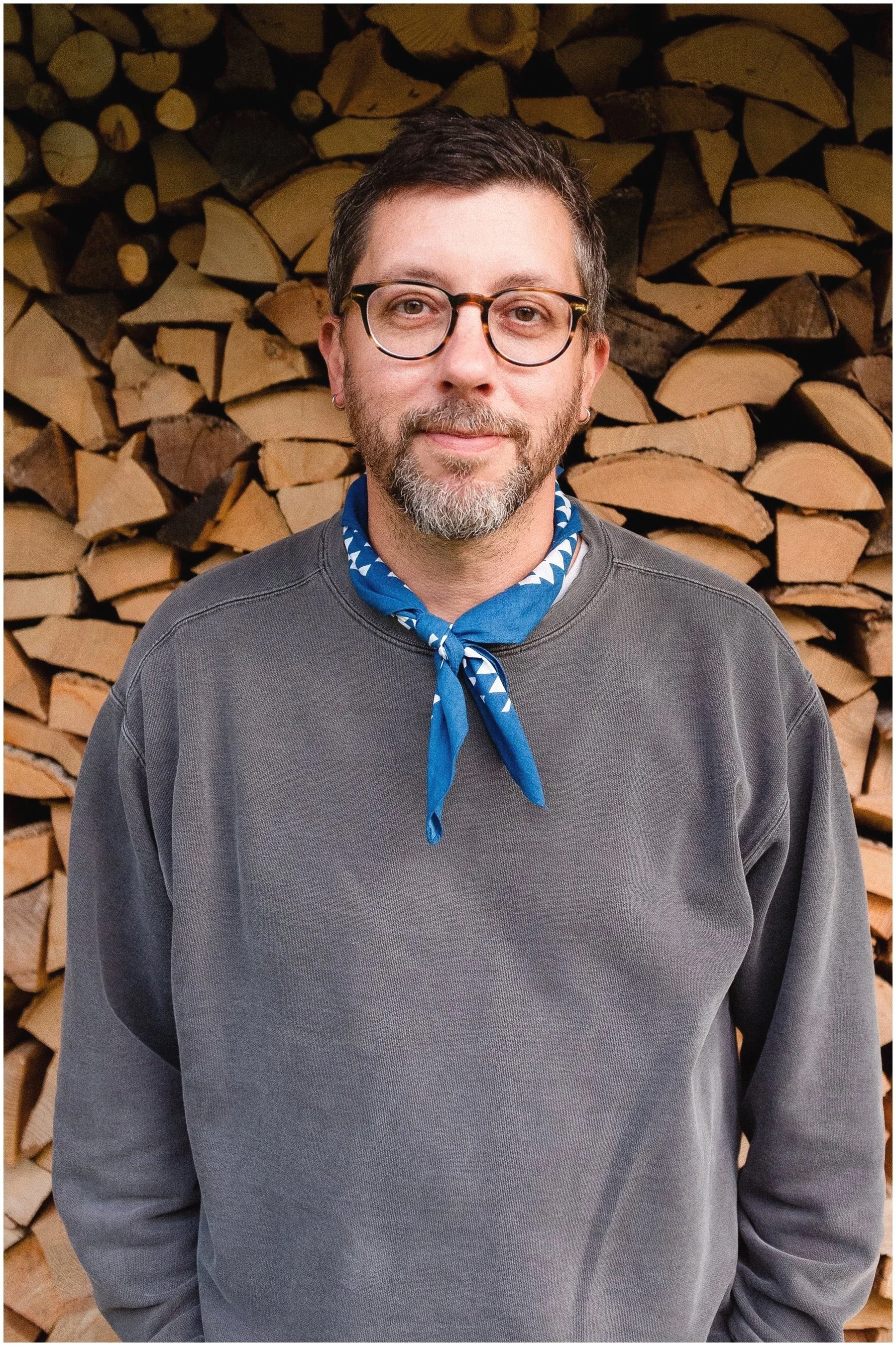 Adam Lowe portrait in front of a pile of logs in Germany. He is wearing a neckerchief and looking stylish