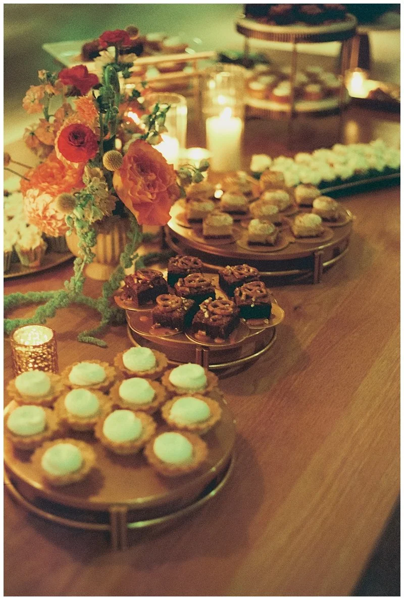 Dessert table with a floral arrangement, candles, and various small sweet treats including cookies and bars on gold trays.