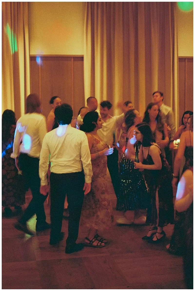 People dancing at a party or event in a dimly lit room with colorful lights and yellow curtains.