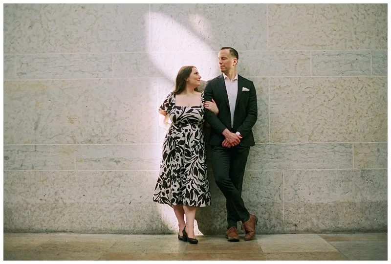 A woman in a patterned dress and a man in a suit standing against a stone wall, looking at each other.
