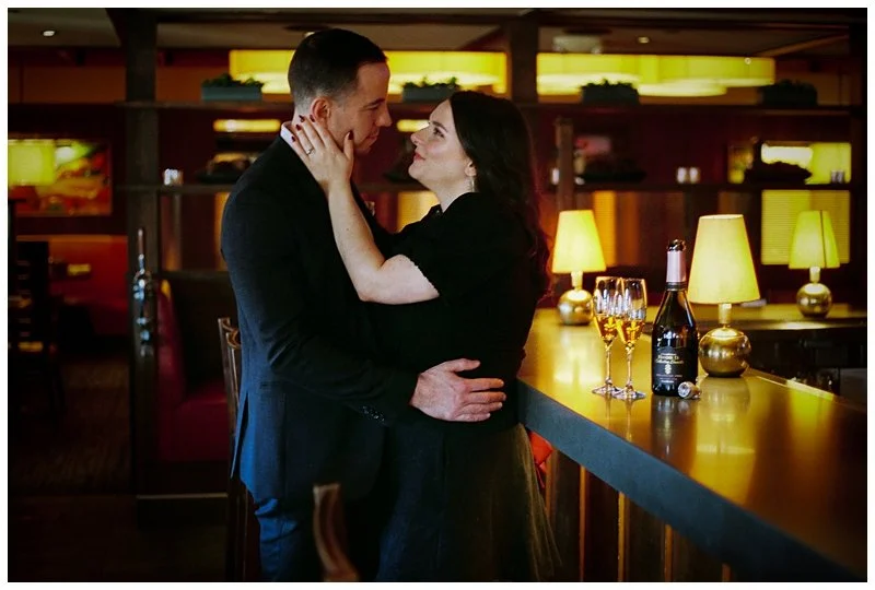 A couple at a bar, gazing into each other's eyes, with a bottle of champagne and glasses on the bar counter, warm lighting, and a cozy atmosphere.