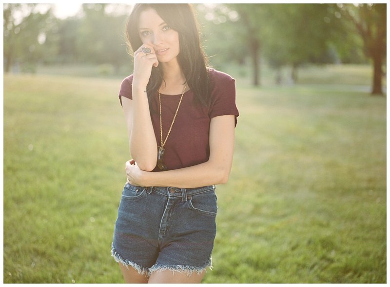A young woman with long dark hair standing outdoors in a park during the daytime, wearing a maroon t-shirt and denim shorts, with sunlight filtering through the trees behind her. Photographed on film