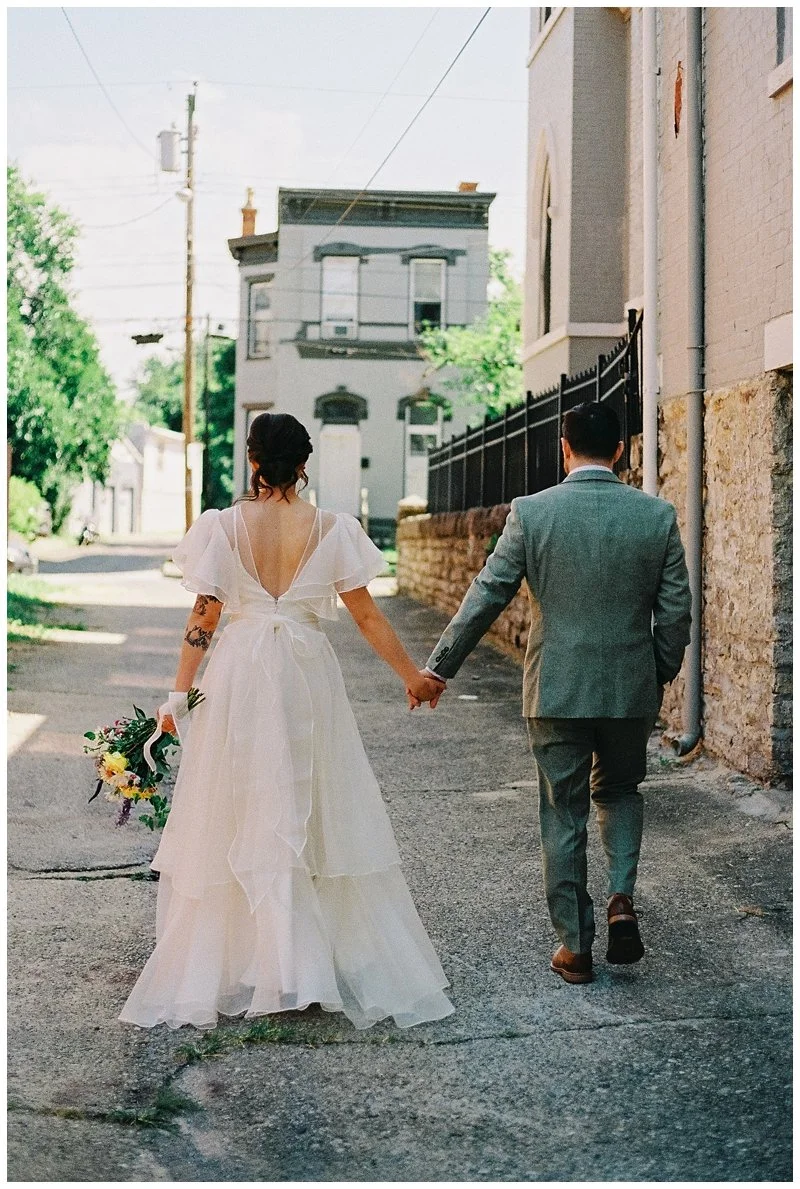 A bride and groom holding hands walk down a street, the bride in a white wedding gown holding a bouquet and the groom in a gray suit. Film Photography
