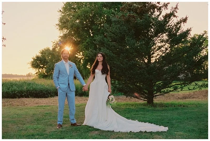 A bride and groom holding hands outdoors during sunset, with a large tree in the background and a grassy field.
