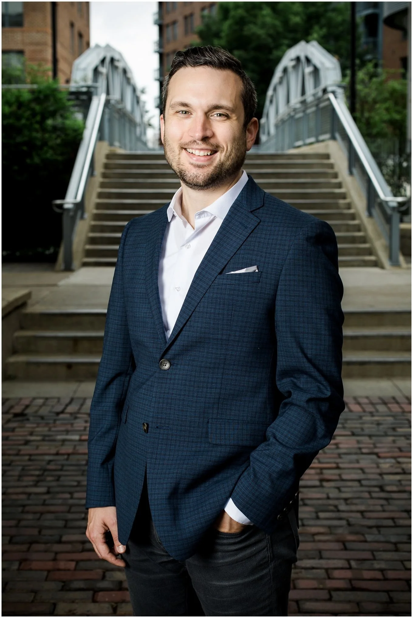 A man in a dark blue checkered suit with a white shirt, smiling, standing outdoors on a brick sidewalk in front of stairs and buildings.