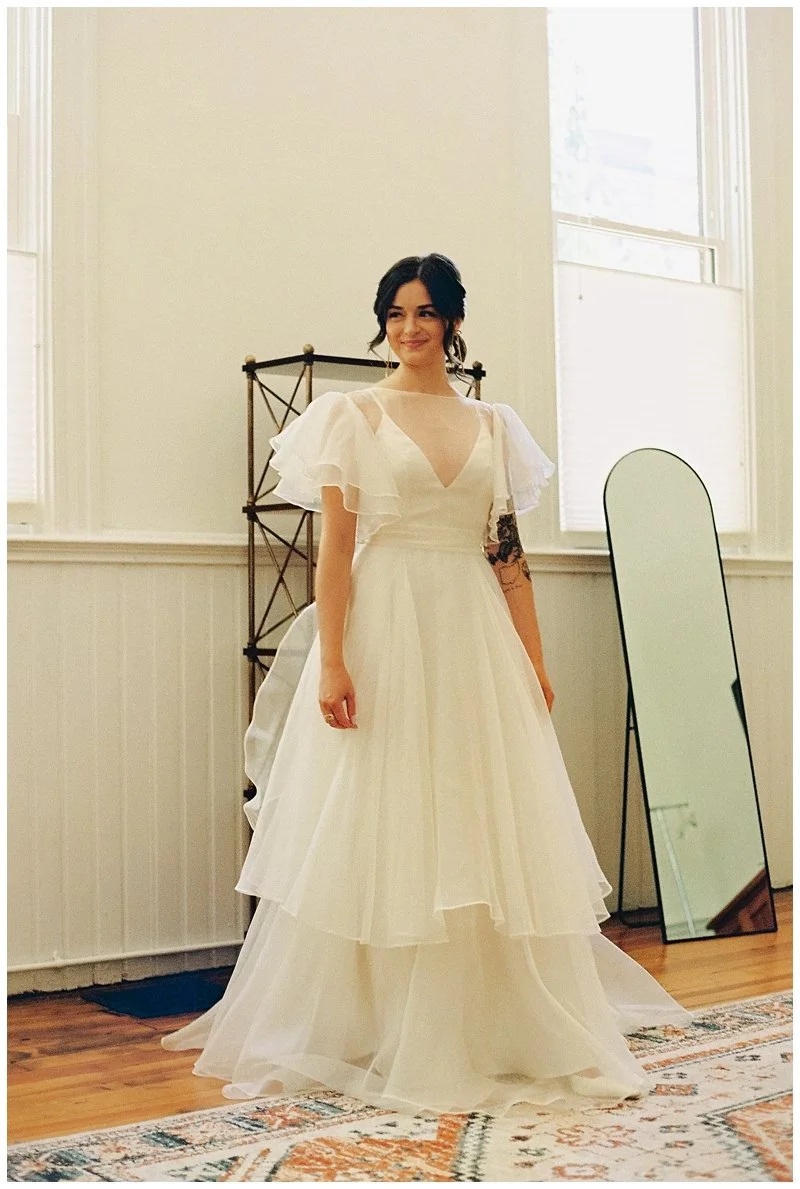 A woman in a white wedding dress with layered tulle skirt and short puffed sleeves stands indoors next to a mirror. She has dark hair styled in loose waves and is smiling.Film Photography