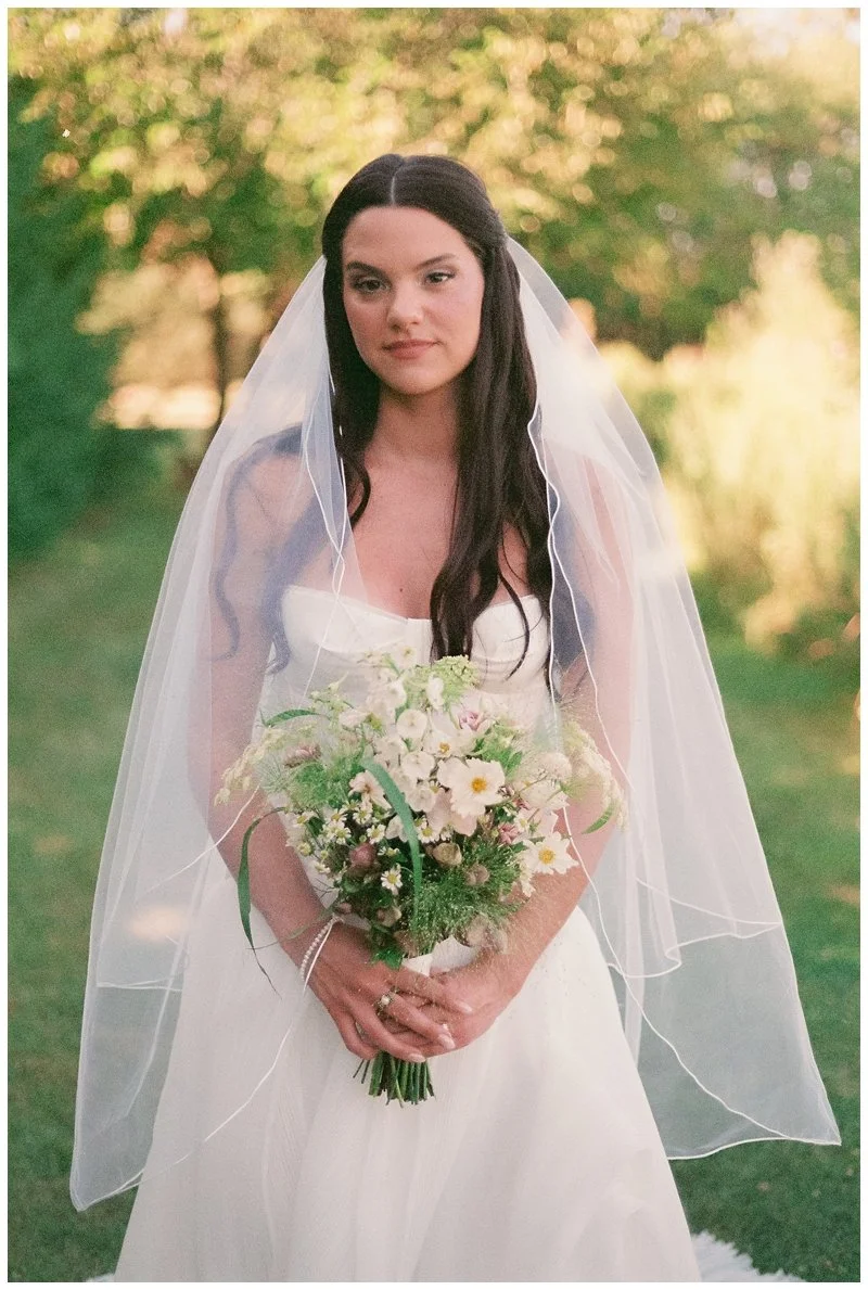 A woman in a wedding dress holding a bouquet of flowers outdoors with trees and green foliage in the background.