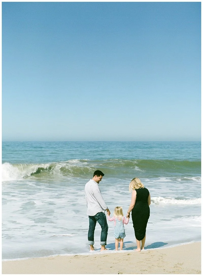 A family of three standing on the beach near the ocean, holding hands, with waves in the background and a clear blue sky. Film Photo