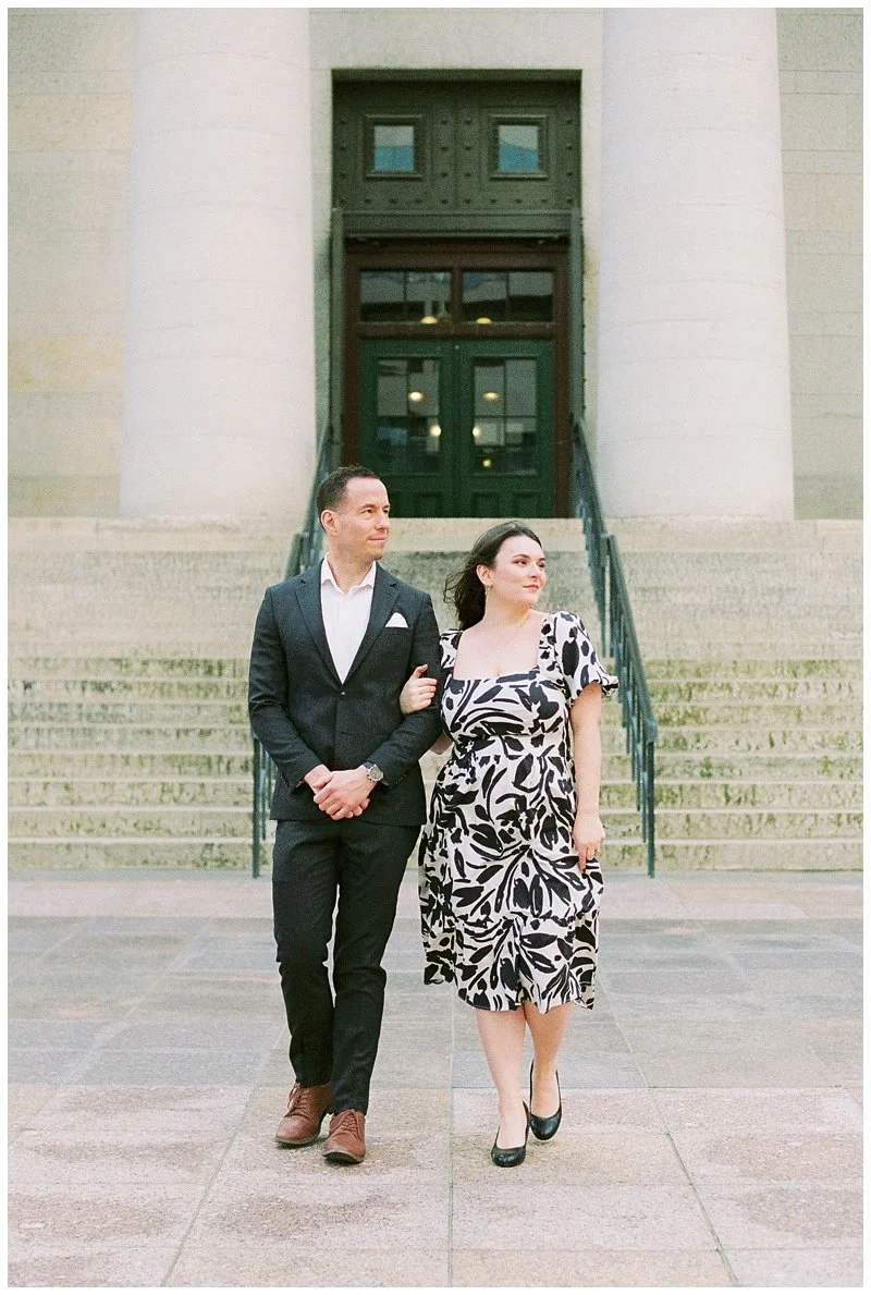 A man and woman walking arm-in-arm in front of a large building with steps and a double door entrance. The man is dressed in a black suit with a white shirt, and the woman is wearing a black and white patterned dress and black heels.