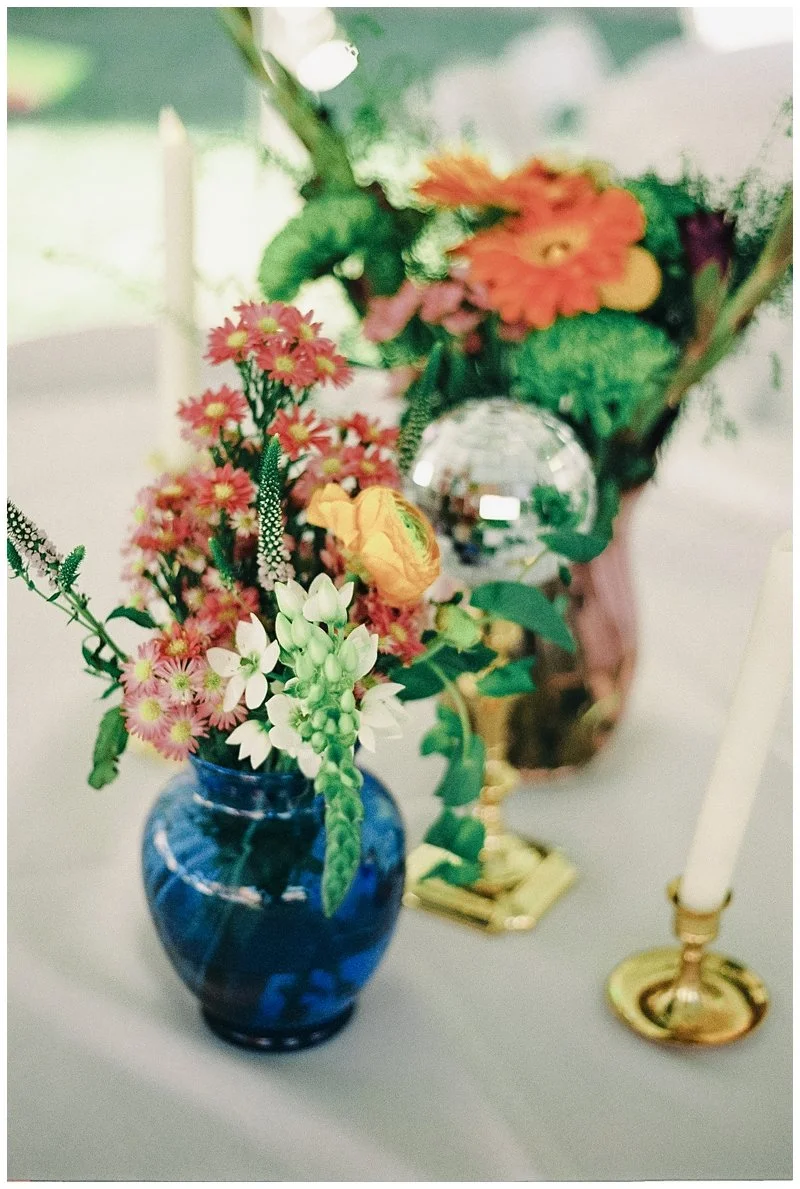 Close-up of a floral arrangement with pink, white, and orange flowers in a blue glass vase, alongside a gold candle holder with a white candle, and a crystal ornament.