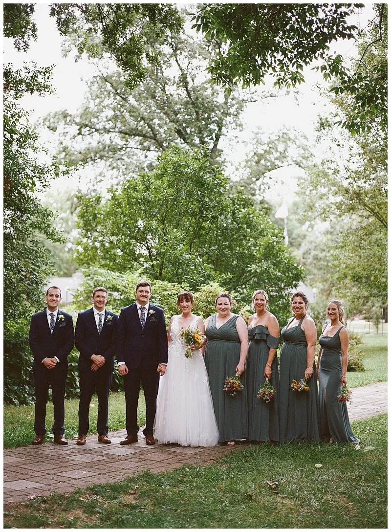 A wedding party outdoors, with three groomsmen, a bride, and four bridesmaids standing on a brick pathway surrounded by trees and greenery.