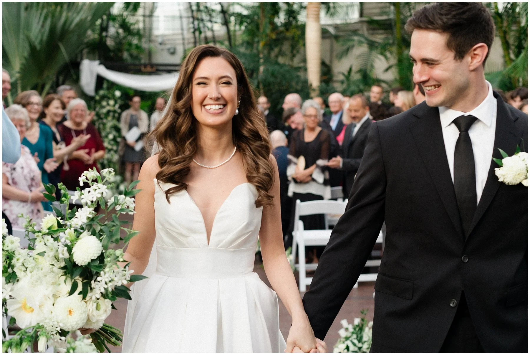 A bride and groom smiling and holding hands during their wedding ceremony, surrounded by guests in an outdoor setting with lush greenery. Franklin Park Conservatory, Adam Lowe Photography