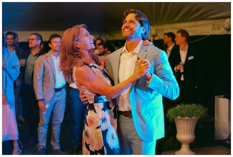 A woman and a man dancing together at a social event or party, smiling and holding hands, with other people in the background.