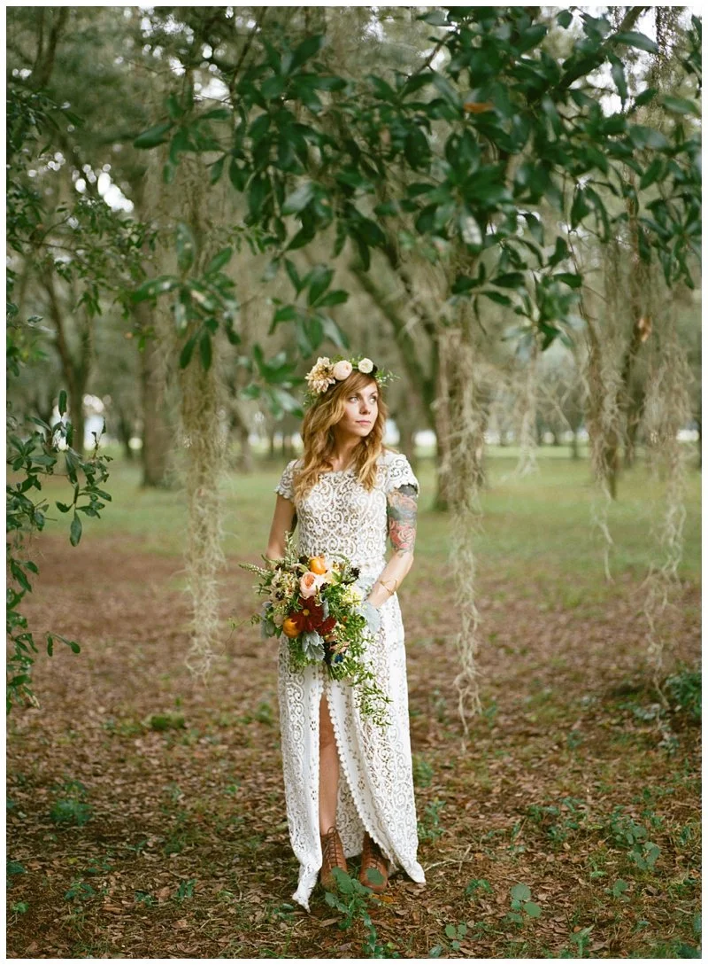 A woman with wavy red hair, wearing a white lace wedding dress with a slit, stands in a forested area. She has a flower crown on her head and is holding a bouquet of flowers, looking off to the side.