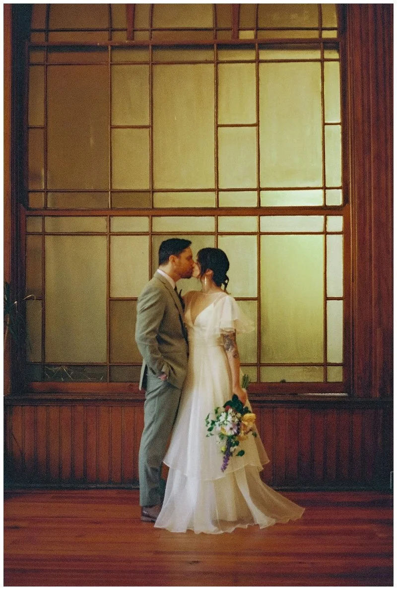 A bride and groom share a kiss indoors, with the bride holding a bouquet of flowers, standing in front of a large wooden window. Film Photography