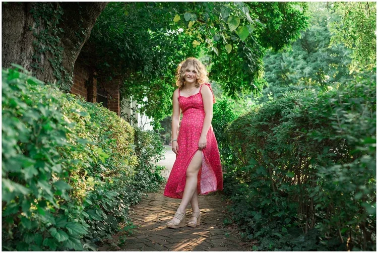 A woman in a pink dress with a high slit standing on a garden path surrounded by lush greenery and trees. Stylish Columbus Ohio graduating senior photo session