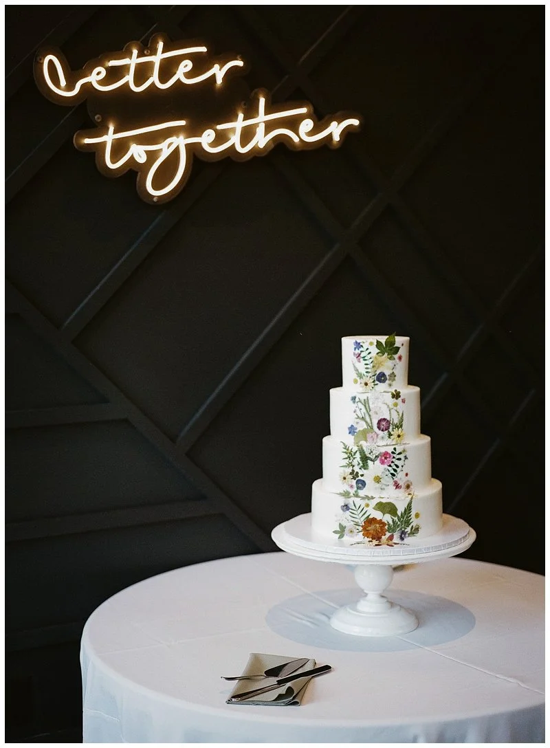 A four-tiered white wedding cake decorated with colorful floral patterns, placed on a white cake stand on a round table covered with a white tablecloth, with a neon sign reading 'better together' in glowing script overhead.