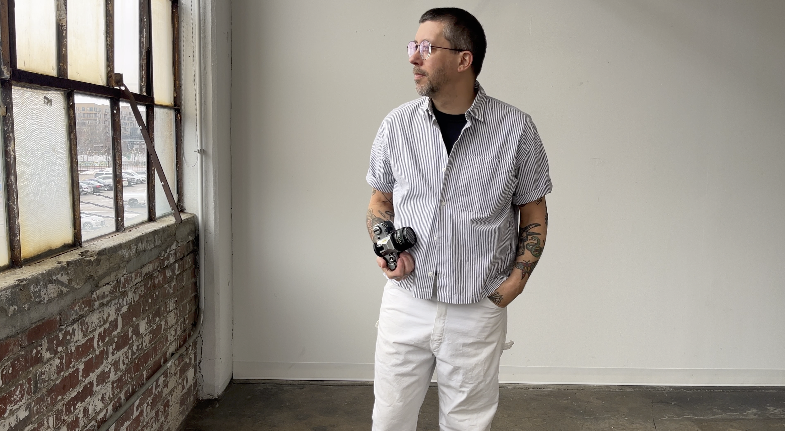 A man with tattoos, glasses, and dark hair stands indoors holding a camera, looking out the window. The room has a brick wall with old windows and a plain white wall.