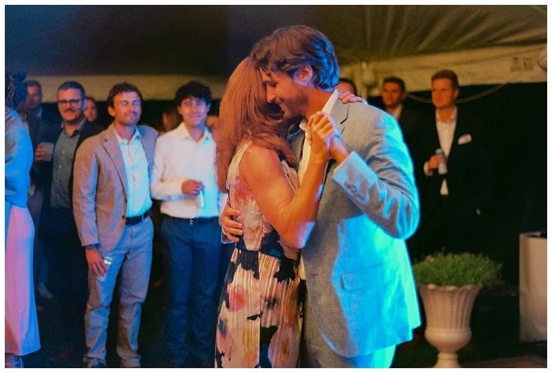 Couple dancing closely at a social event, with onlookers dressed in business attire in the background.