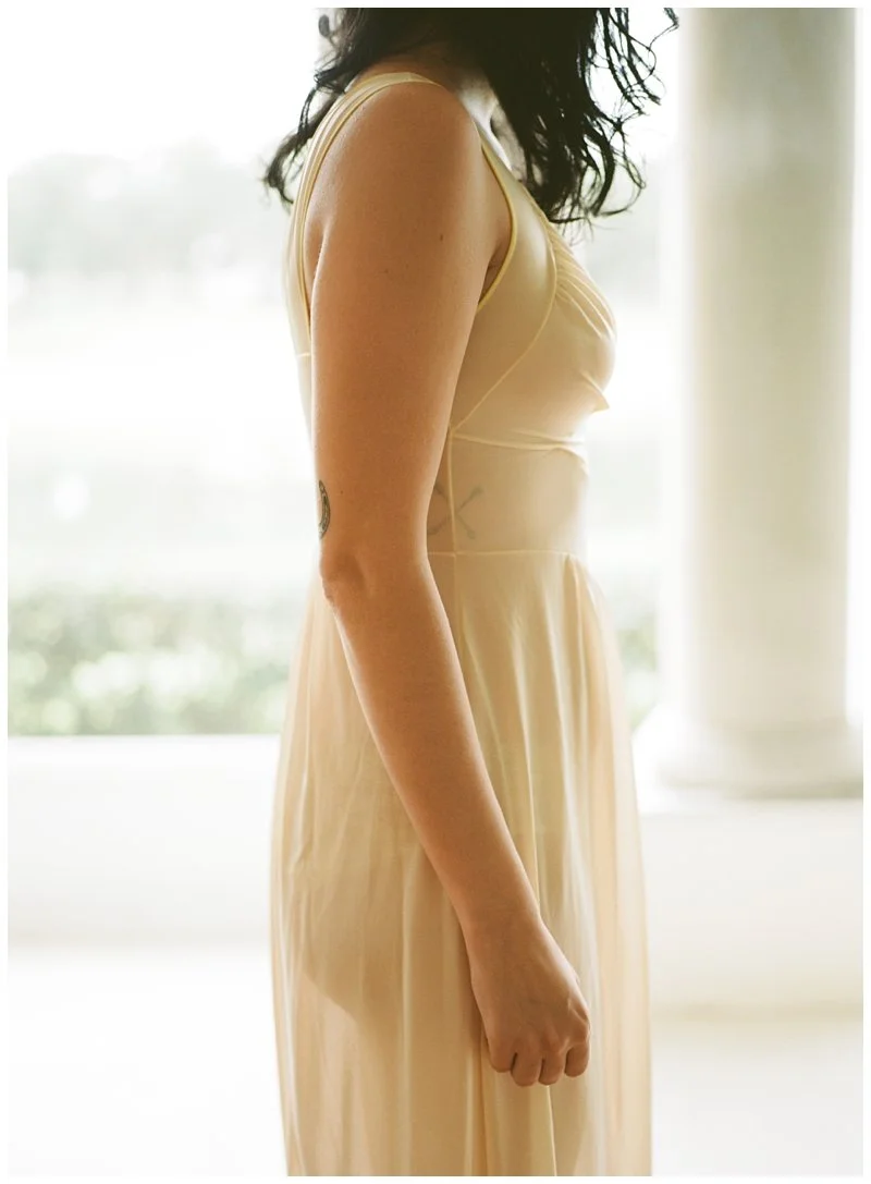 A woman in a sleeveless, light-colored dress standing indoors near a window with natural light. Adam shot this on film