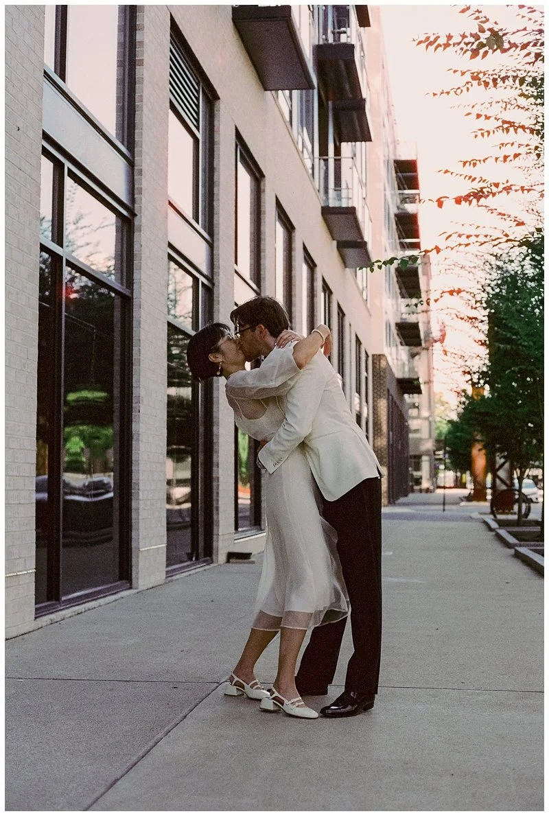 A couple dressed in vintage wedding attire sharing a kiss on a city sidewalk during sunset, with apartment buildings and trees in the background.