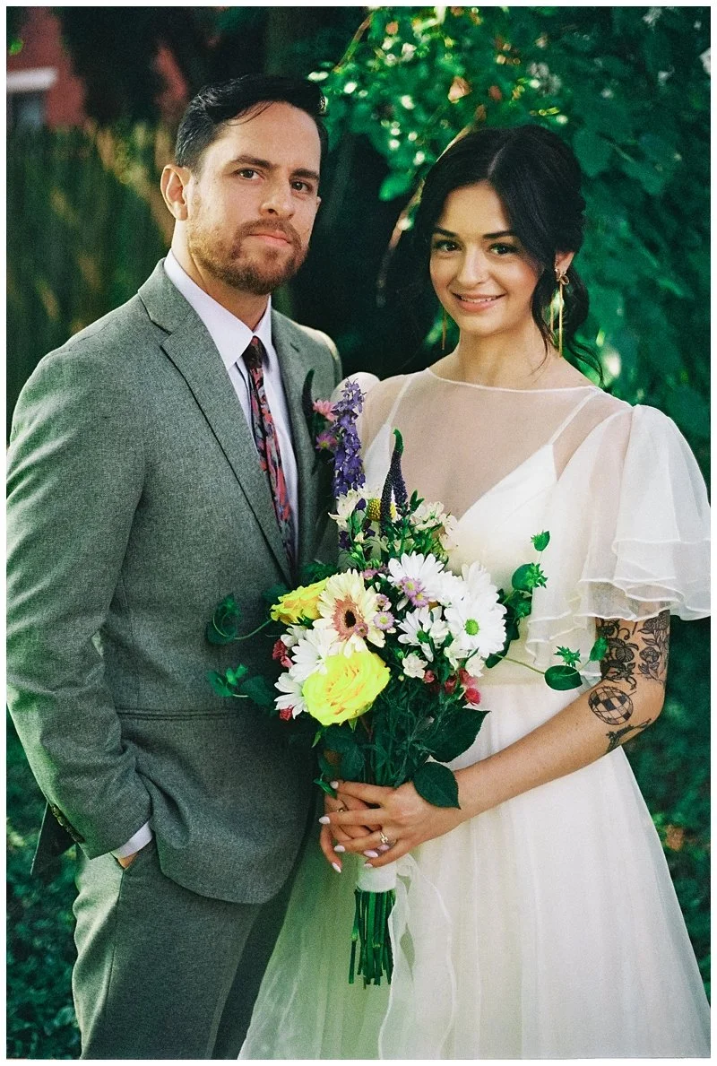 A man and woman, dressed in formal attire, standing outdoors with green foliage in the background. The woman is holding a bouquet of colorful flowers and has a tattoo on her arm. They are smiling at the camera.