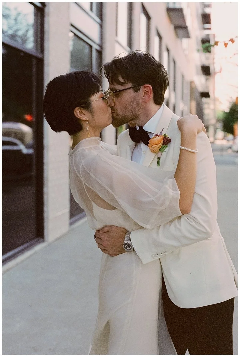 A wedding couple kissing outdoors, with the woman in a white dress and the man in a white tuxedo with a black bow tie and boutonniere, embracing each other on a city sidewalk.