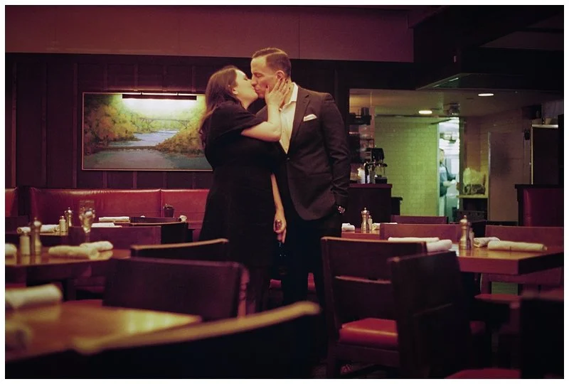A couple sharing a kiss in a dimly lit restaurant or cafe, with tables and chairs in the foreground and a landscape painting on the wall in the background.