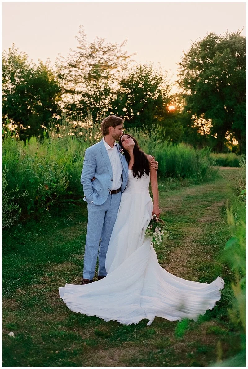 A bride and groom in wedding attire standing outdoors at sunset, surrounded by greenery and trees, sharing an intimate moment.