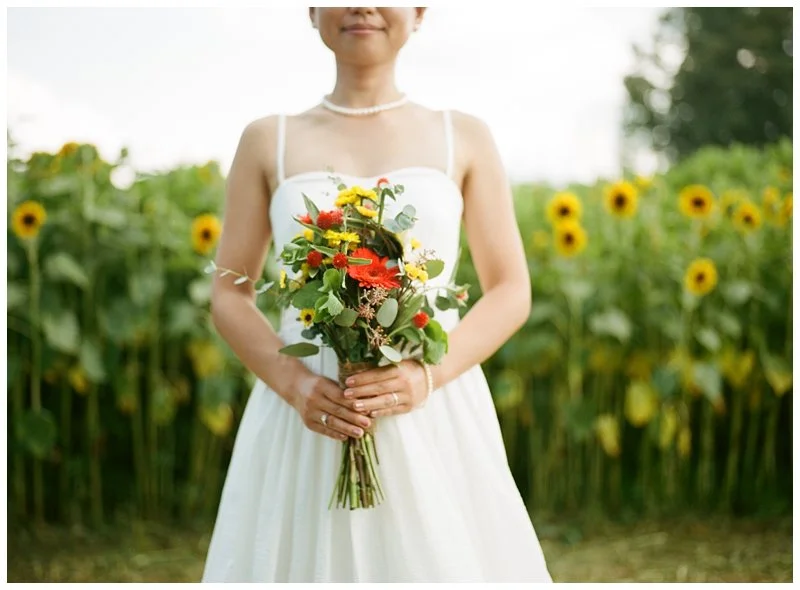 A woman in a white dress holding a colorful bouquet of flowers stands in front of a sunflower field. Shot on film