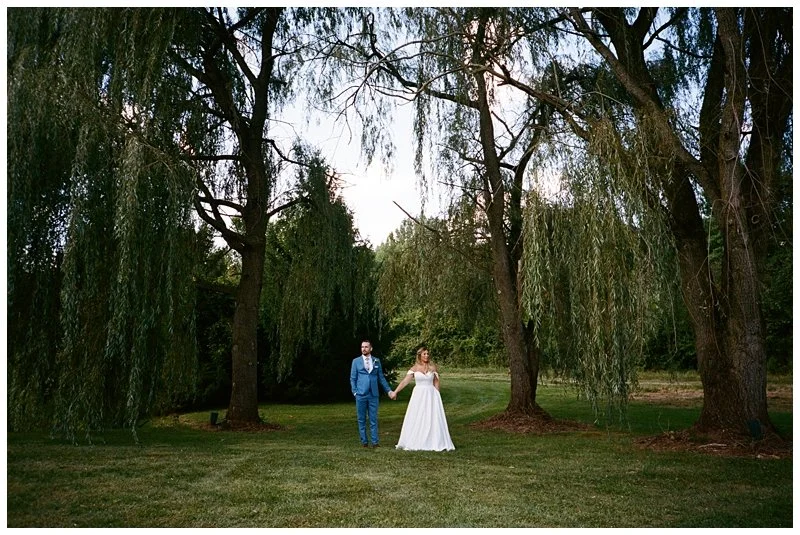 A bride and groom holding hands in a grassy park with tall trees and hanging branches.