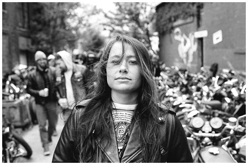 A young woman with long hair and a leather jacket standing in front of a row of motorcycles on a city street.