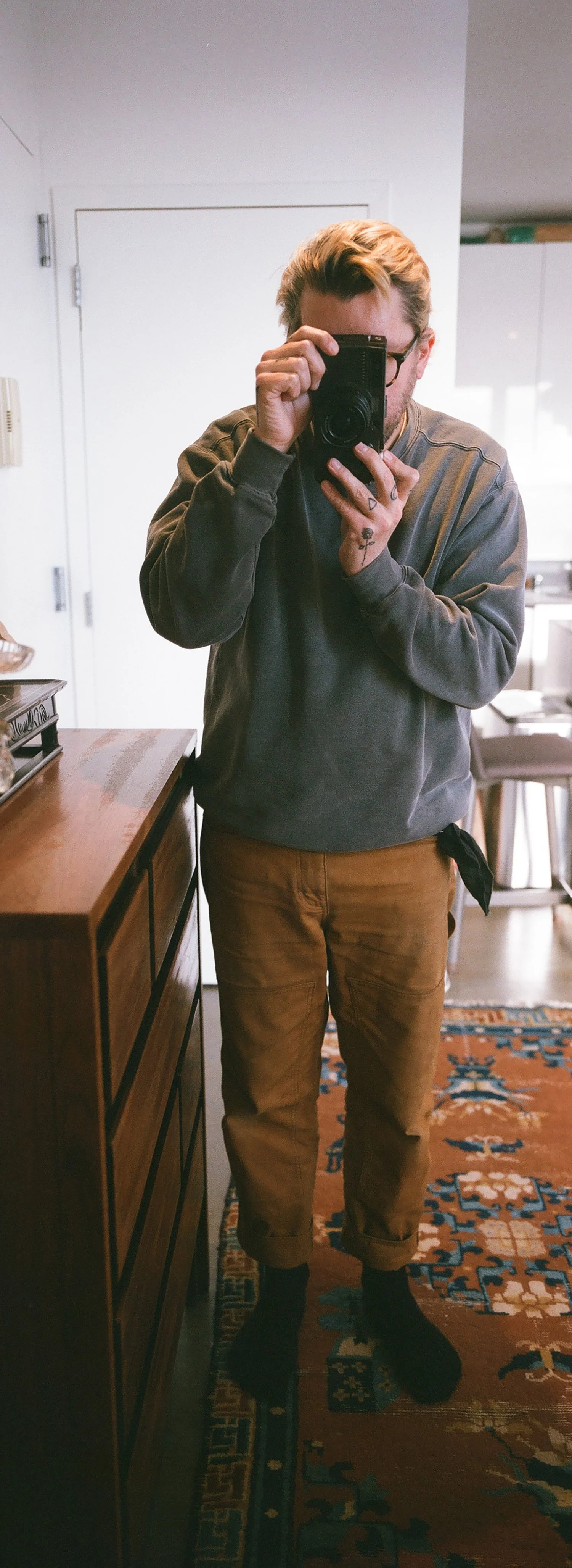 A person taking a mirror selfie with a camera. The individual has light brown hair, glasses, a dark gray sweatshirt, and light brown pants rolled up at the cuffs, standing indoors in front of a white door.