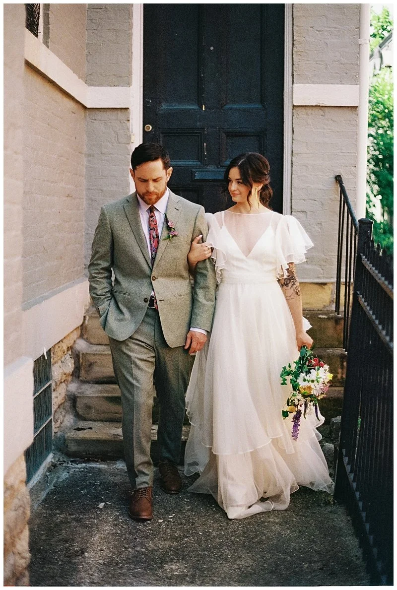 A bride and groom walking together, with the bride supporting the groom's arm. The bride is holding a bouquet of flowers and wearing a white wedding dress with sheer sleeves. The groom is dressed in a gray suit with a floral tie. They are walking dow