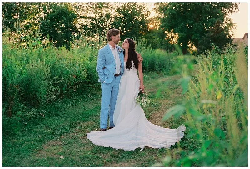 A bride and groom are standing on a grassy path in a lush green field at sunset, gazing into each other's eyes. The bride is wearing a white wedding gown and holding a small bouquet, while the groom is dressed in a light blue suit.
