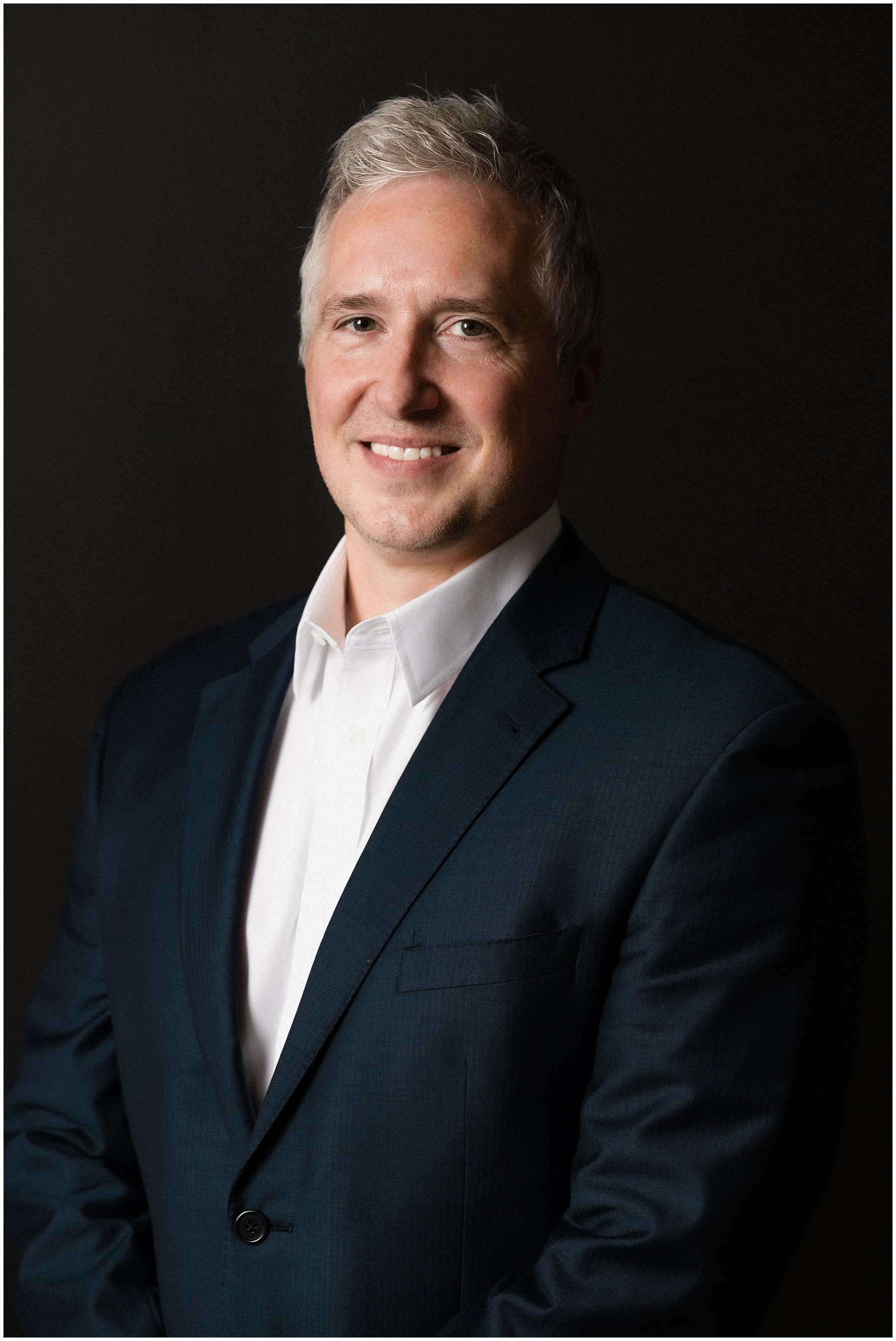 A man with gray hair wearing a dark suit and white shirt, smiling against a black background.
