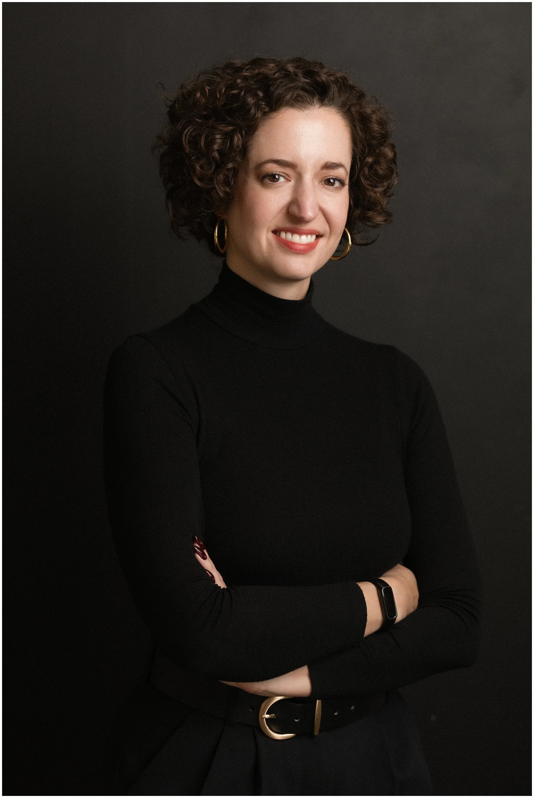 A woman with short, curly brown hair, wearing a black turtleneck, gold hoop earrings, and a black watch, standing against a dark background with her arms crossed and smiling at the camera.
